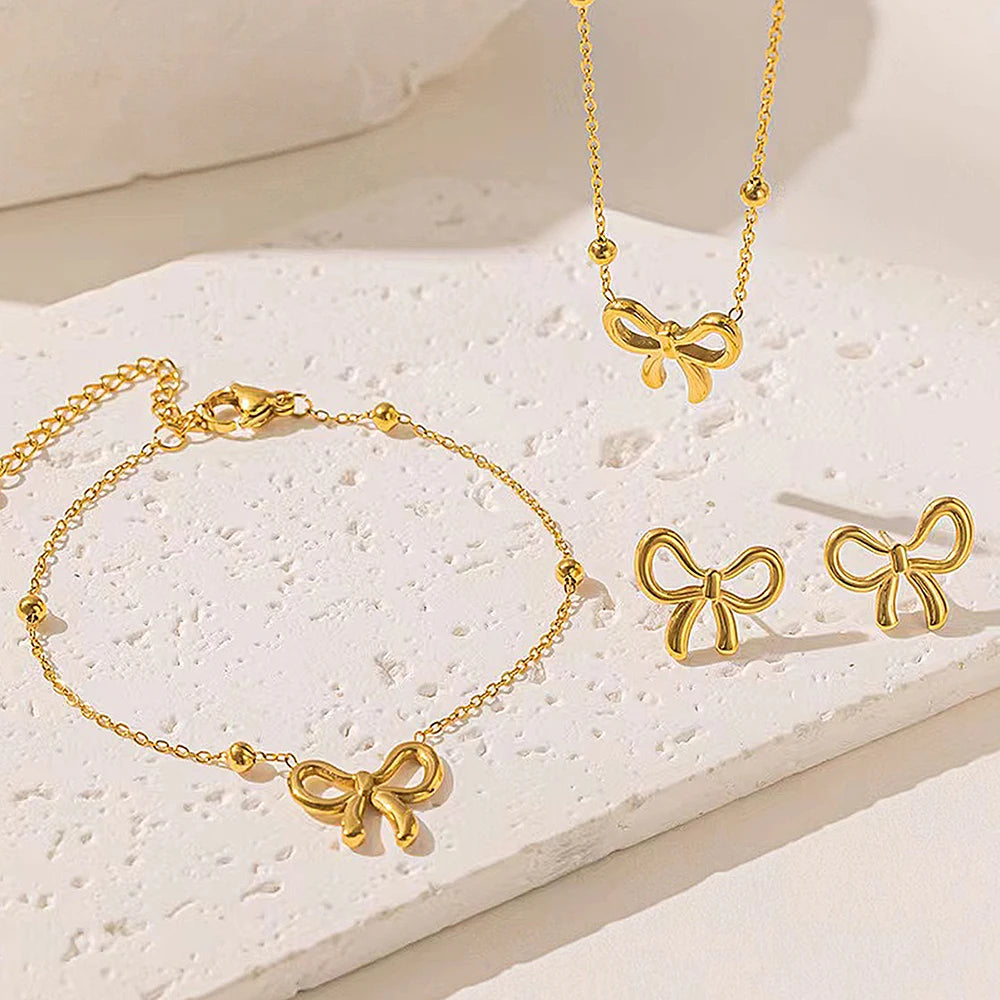 Gold bow-shaped jewelry set including necklace, bracelet, and earrings on a light stone surface.