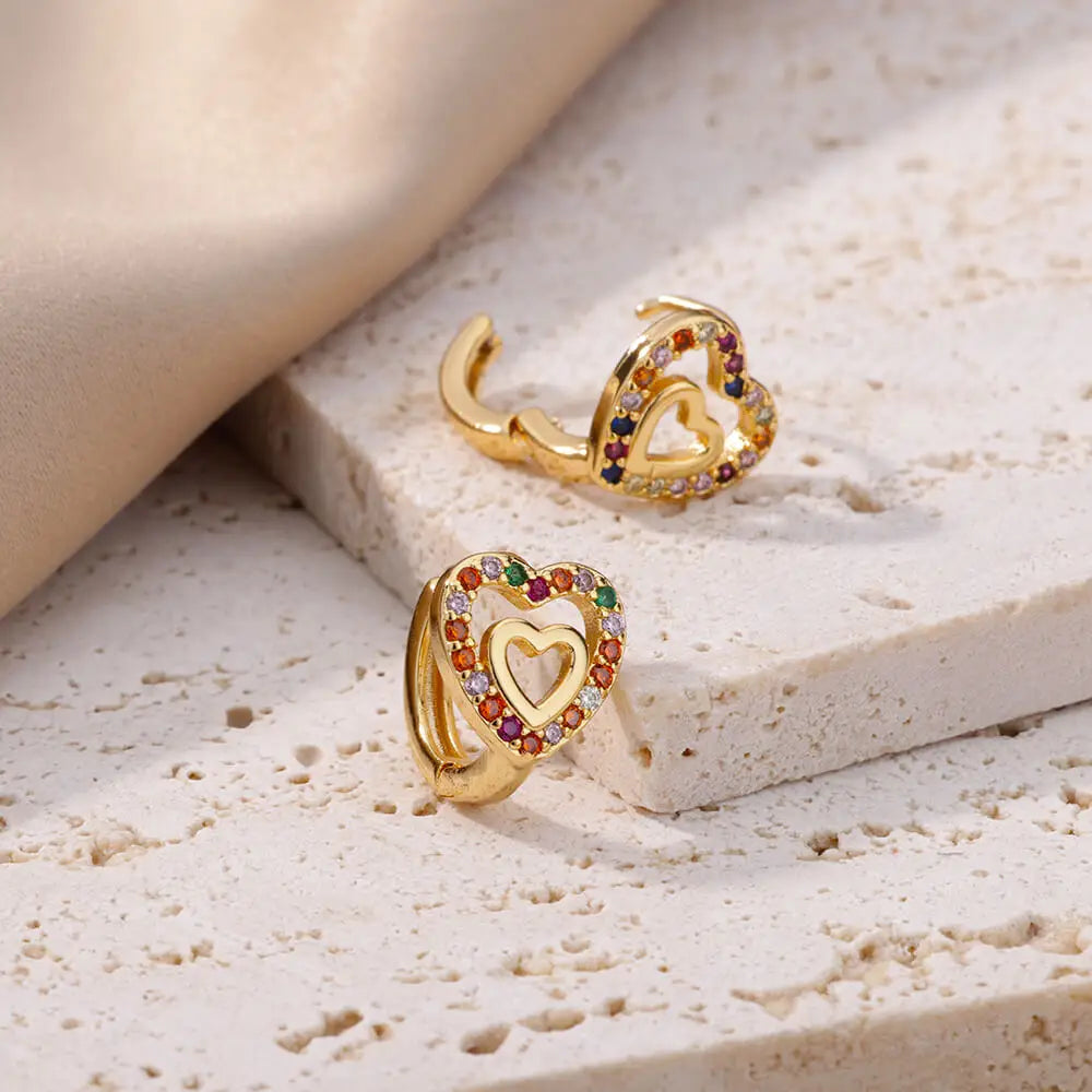 Gold heart-shaped earrings with colorful stones on a textured beige surface