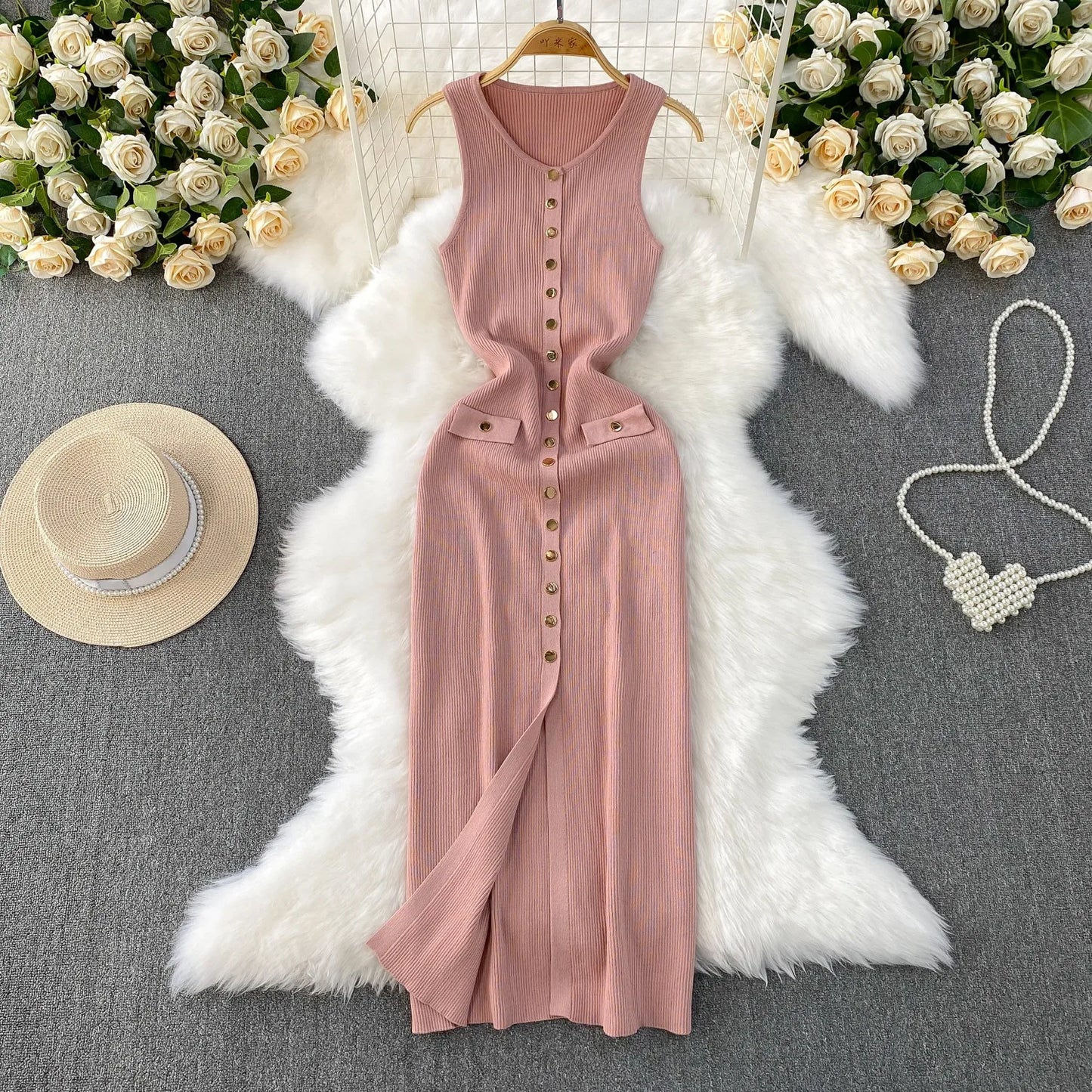 Pink sleeveless dress on a hanger with decorative elements like flowers, a hat, and necklace on a gray surface.