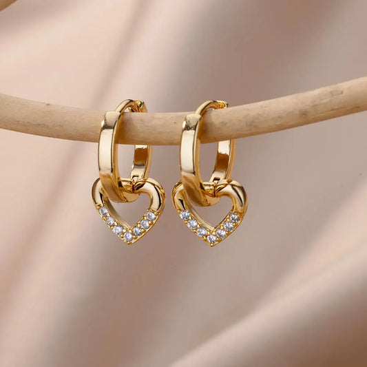 Gold heart-shaped hoop earrings with embedded stones on a wooden stick against a soft pink background
