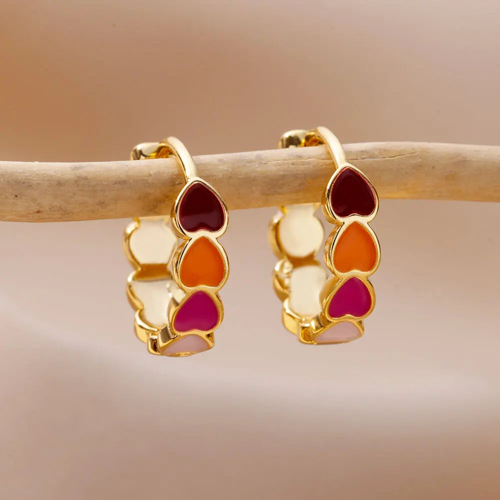 Gold earrings with colorful heart-shaped stones on a wooden stick against a beige background
